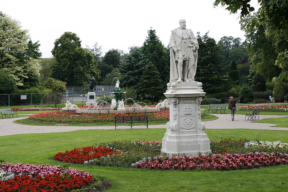 The image shows Beacon Park in Lichfield. There is a fountain and many flowerbeds in the park, as well as a statue of Edward VII.